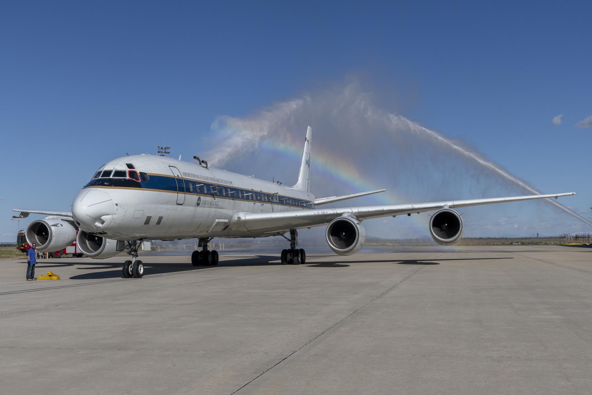 An aircraft taxis through a water salute streaming from water hoses from two firetrucks positioned on either side of the aircraft. The converging water streams create a rainbow above the aircraft as it passes under the arc of spraying water.